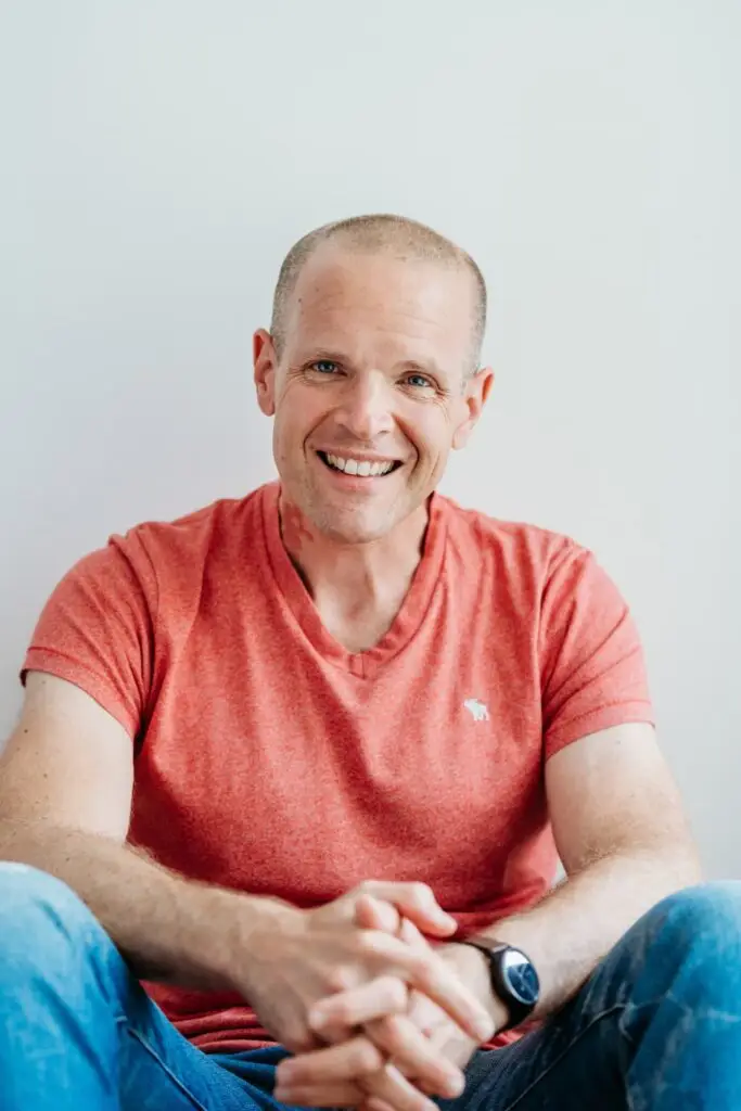Author of Everyday WEightloss, 365 tips to lose weight and keep it off, Neil Roberto Williams. He is sat on the floor leaning against a white wall wearing a pink tshirt and blue jeans with his arms out in front resting on his legs